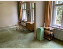 Ein schlicht eingerichteter Raum mit grünen Teppichboden, einer Rattan-Stuhl, einem kleinen Holzschrank und einem Regal mit grüner Säule. Zwei große Fenster mit beigen Vorhängen lassen Tageslicht herein, mit Blick auf Bäume draußen.