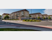 Mehrstöckige Wohngebäude mit hellgelben Fassaden und Satteldächern umgeben einen Kreisverkehr, der mit Pflanzen und Blumen gestaltet ist. Im Erdgeschoss eines Gebäudes befinden sich Geschäfte, während auf der Straße geparkte Autos und Gehwege mit Bordstein und Pflastersteinen sichtbar sind.