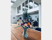 Moderne Küche mit hohen weißen Wänden, großen Fenstern und Oberlichtern, die viel Tageslicht einlassen. Im Vordergrund steht ein Holztisch mit einer Glasvase, die künstliche Blumen enthält, und im Hintergrund befinden sich eine Kücheninsel aus Edelstahl sowie ein großer Kaktus als Dekoration.