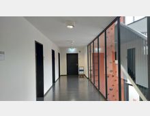 Flur in einem modernen Gebäude mit schwarzen Türen auf der linken Seite und einer Glaswand rechts, hinter der ein roter Bereich mit einer Kletterwand sichtbar ist. Am Ende des Flurs führt eine Tür mit einem Notausgangsschild darüber hinaus.