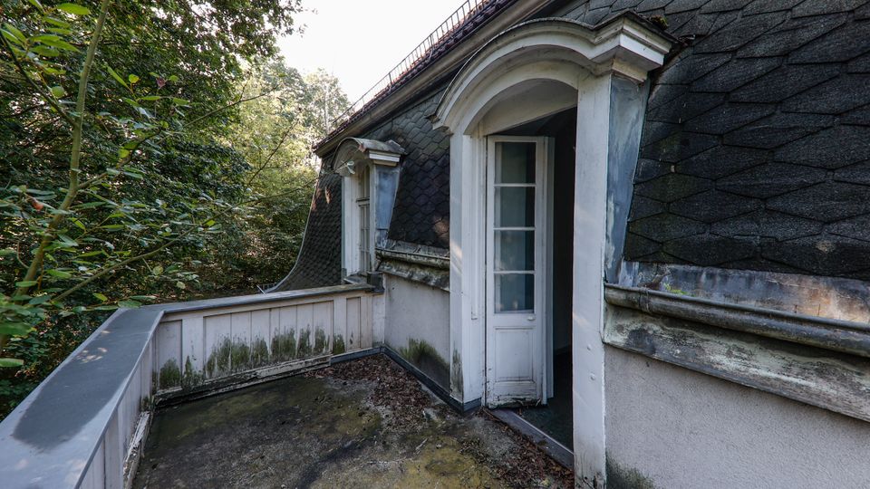 Kleiner Dachbalkon an einem älteren Gebäude mit dunkel gedecktem Mansarddach und Rundbogen-Dachgaube mit geöffnetem Türfenster. Der Betonboden und die Brüstung sind verwittert und von Moosspuren bedeckt, umgeben von dichtem Wald.