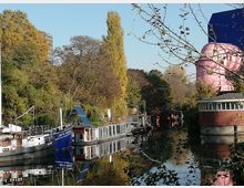 Kanalufer mit mehreren Hausbooten und kleinen Booten, umgeben von herbstlichen Bäumen. Im Hintergrund ein rosafarbenes, kuppelähnliches Gebäude und ein modernes, blaues Gebäude.