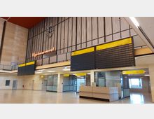 Innenansicht eines Flughafengebäudes mit großer Abflug-Anzeigetafel, einer Uhr und einem Neonschild mit der Aufschrift „Restaurant“. Der Bereich ist modern gestaltet mit hellen Wänden, hohen Decken und mehreren gelben Wegweisern für Abflüge.