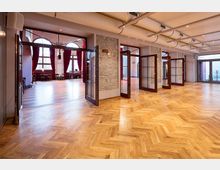 Innenansicht eines großen Veranstaltungsraums mit hellem Fischgrätparkett, Glas-Doppeltüren und freiliegenden Lüftungsrohren an der Decke. Im Hintergrund sind hohe Fenster mit roten Vorhängen und eine weitere Halle mit Holztischen und Stühlen sichtbar.