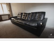 Ein schlicht eingerichteter Raum mit Teppichboden und einer schwarzen Ledercouch mit Abnutzungsspuren. Im Hintergrund befindet sich ein großes, einfach verglastes Fenster mit Heizkörper darunter.