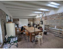 Ein großer Raum mit sichtbaren Backsteinwänden und einer weißen Holzbalkendecke. Im Raum stehen ein großer Holztisch mit Stühlen, ein Ofen, ein Kühlschrank und mehrere Stühle, Sofas und Haushaltsgegenstände; Oberlichter lassen Tageslicht einfallen.