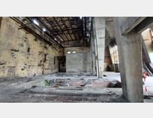 Innenansicht einer verlassenen Industriehalle mit hohen Wänden aus hellen Ziegelsteinen und großen Betonstützen. Der Raum zeigt abgenutzte Strukturen, offene Schächte im Boden und eine teilweise beschädigte Decke mit sichtbaren Metallträgern.