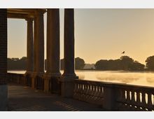 Blick von einer Säulenarkade am Ufer auf einen ruhigen See im Morgenlicht, mit Nebel über der Wasseroberfläche und silhouettierten Bäumen im Hintergrund. Eine Brüstung mit Balustern verläuft entlang der Arkade, und ein Vogel fliegt über den See.
