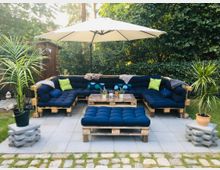 Terrasse mit Sitzmöbeln aus Paletten, ausgestattet mit dunkelblauen Polsterkissen und grünen Dekokissen. Ein zentraler Couchtisch sowie ein großer Sonnenschirm bieten ein gemütliches Ambiente; umgeben von Pflanzen, Rasen und dichten Büschen.