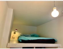 Kleiner Schlafbereich auf einer erhöhten Plattform in einem schlichten Raum mit hellen Wänden, einem Bett mit türkisem Bezug, einer Wandlampe mit rotem Schirm und einer hängenden Glühbirne als Beleuchtung.