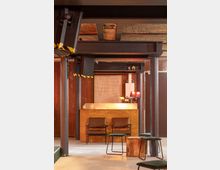 Innenraum eines modernen Raumes mit industriellem Stil, geprägt von sichtbaren Stahlträgern, Holzwänden und einer Holztheke. Es gibt Stühle, Hocker und eine Tischlampe, beleuchtet von warmer Beleuchtung.