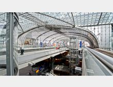 Modernes Bahnhofsterminal mit gläserner Dachkonstruktion und mehreren Ebenen. Sichtbar sind ein transparenter Aufzug, Bahnsteige, Geschäfte im unteren Bereich sowie Fußgänger auf der Galerie.