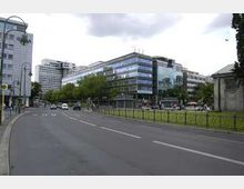 Straßenszene in einer Stadt mit modernen Bürogebäuden, großen Fenstern und mehreren Etagen. Im Vordergrund verläuft eine mehrspurige, asphaltierte Straße, daneben eine grüne Rasenfläche mit einem Gehweg und einigen Fußgängern.