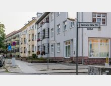 Straßenecke mit mehrstöckigen Wohngebäuden, die Balkone mit Blumen geschmückt haben, und einem Ladenfenster mit der Aufschrift "Physiotherapie". Straßenschilder markieren die "Heinrich-Zille-Str." und Gehwege sind mit Kopfsteinpflaster ausgelegt.