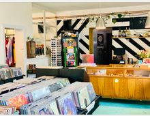 Ein Plattenladen mit Holztheke im Vordergrund, dahinter schwarz-weiß gestreifte Wände. Schallplatten sind in Regalen sortiert, während im Hintergrund auch Accessoires wie Sonnenbrillen und Kleidung sichtbar sind. Hängeleuchten und Pflanzen schmücken den Raum.