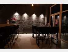 Dunkel beleuchtetes Café oder Restaurant mit modernen schwarzen Barhockern an einer Theke links und hohen Tischen mit Stühlen rechts. Die Wand besteht aus grobem Putz, der durch punktuelle Strahler beleuchtet wird, und rechts ist eine große Glasfront sichtbar.