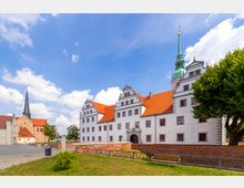Weitläufiger Platz mit einem historischen Gebäude mit weißer Fassade, roten Dachziegeln und kunstvoll verzierten Giebeln; daneben eine Kirche mit spitzem, schwarzem Turm. Im Vordergrund befinden sich eine Grünfläche und Sitzbänke, im Hintergrund ein blauer Himmel mit Wolken.