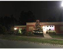 Ein einstöckiges Wohnhaus mit flachem Dach, roter Fassade und weißer Eingangstür, dekoriert mit weihnachtlicher Beleuchtung. Vor dem Haus steht ein großer Baum mit Lichterketten, umgeben von weiteren beleuchteten Pflanzen; im Vordergrund befindet sich eine beleuchtete Straße mit Kopfsteinpflasterstreifen.
