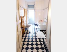 Helles Badezimmer mit schwarz-weiß kariertem Fliesenboden, weißen Wandfliesen, einer Badewanne rechts, einem hölzernen Regal mit Handtüchern links und einer Glastür mit Milchglasfenster im Hintergrund.