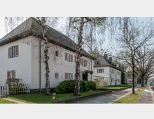 Straßenszene mit weißen Einfamilienhäusern im traditionellen Stil, die Schrägdächer und Fensterläden aufweisen. Im Vordergrund stehen Birken und andere Bäume, die Straße ist mit Gehweg und Bordstein ausgestattet, und es sind vereinzelt geparkte Autos zu sehen.