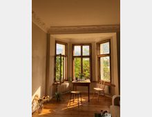 Wohnraum mit einem Erker, der durch drei hohe Holzfenster mit Blick auf grüne Bäume geprägt ist. Der Raum hat einen Holzboden, Stuckverzierungen an der Decke und einfache Möbel, darunter ein Holztisch mit Stühlen.