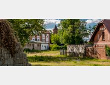 Grüne Freifläche mit vereinzelten Pflanzen und Büschen, umgeben von Gebäuden aus rotem Backstein und hellgrauem Beton; im Hintergrund sind ein Kirchturm und weitere Bäume zu sehen. Ein Baum und eine überwucherte Mauer im Vordergrund rahmen die Szenerie ein.