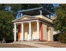 Gelbes Pavillongebäude im klassizistischen Stil mit vier weißen Säulen und rot abgesetzten Wänden, flankiert von Bänken und umgeben von Bäumen. Das Dach ist von einem blau-weiß gestreiften Sonnendach bedeckt.