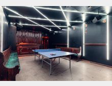 Innenraum eines dunklen Freizeitraums mit Tischtennisplatte in der Mitte, Holzbänken an den Seiten und einer Holztheke an der Rückwand. Decke und Wände sind mit hellen LED-Lichtstreifen in geometrischen Linienmustern und vertikalen Lichtleisten akzentuiert.
