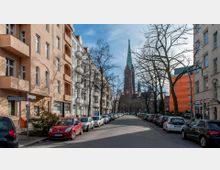 Straßenansicht in einer Stadt mit mehrstöckigen Wohngebäuden auf beiden Seiten, parkenden Autos am Straßenrand und einer großen Backsteinkirche mit spitzem grünen Turm im Hintergrund. Kahle Bäume säumen die Straße bei hellem Tageslicht.