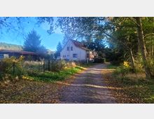 Ein ländliches Wohnhaus mit grauer Fassade und Satteldach steht am Ende eines gepflasterten Weges, umgeben von Bäumen, Sträuchern und Zäunen. Links im Bild befinden sich ein kleiner Garten und ein Nebengebäude mit rotem Dach.