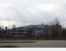 Eine große, rechteckige Industriehalle mit grauer Fassade, umgeben von kahlen Bäumen und niedrigem Gestrüpp, bei bewölktem Himmel und Sonnenstrahlen, die durch die Wolkendecke fallen. Im Vordergrund befindet sich ein leerer, gepflasterter Parkplatz.