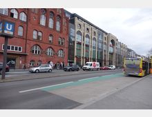 Straßenszene mit einer roten Backsteinfassade eines historischen Gebäudes links und einem modernen Einkaufszentrum mit großen Glasfronten rechts. Im Vordergrund fahren Autos und ein gelber Linienbus auf einer mehrspurigen Straße mit grün markiertem Fahrradstreifen; ein U-Bahn-Schild weist auf eine nahegelegene Station hin.