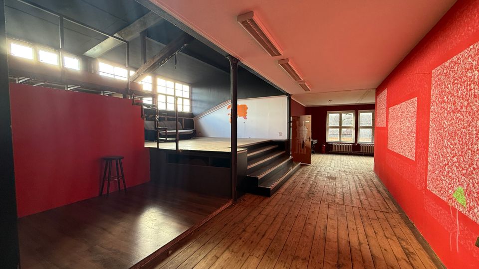 Innenraum einer ehemaligen Industriehalle mit breitem Holzfußboden, roten Wänden und einer erhöhten Plattform mit Treppe. Große Fensterreihen an der linken Seite und am Ende des Raums lassen viel Tageslicht hinein; dunkle Stahlträger und eine hohe Decke prägen die Architektur.