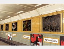 U-Bahn-Station mit der Beschilderung „Hermannplatz“, gelben Wandfliesen und mehreren Kunstwerken in dunklen Farbtönen an der Wand. Unterhalb der Kunstwerke befinden sich Werbeplakate und Stationsschilder auf grün gekacheltem Hintergrund.
