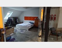 Ein Schlafzimmer in einem Dachgeschoss mit schräger Decke und einem Dachfenster. Im Raum befinden sich ein weißes Bett, ein oranger Wandpolster, eine Kommode, ein offener Kleiderständer sowie ein Spiegel; der Boden ist aus Beton.