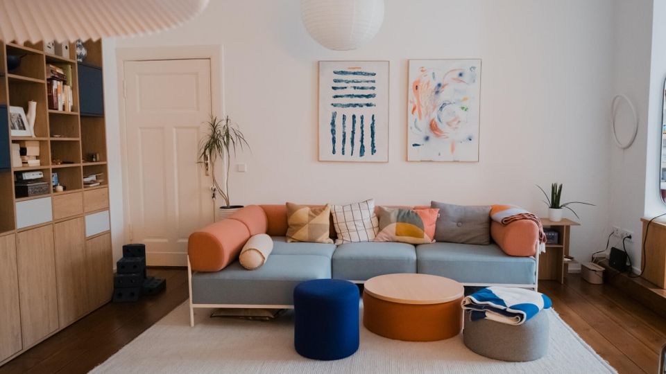 Bright modern living room with white walls, a light blue sectional sofa with colorful cushions, and round ottomans and a low coffee table on a white rug over dark wood floors. Built-in wooden shelving, a white door, potted plants, and two framed abstract artworks hang beneath a round pendant light.