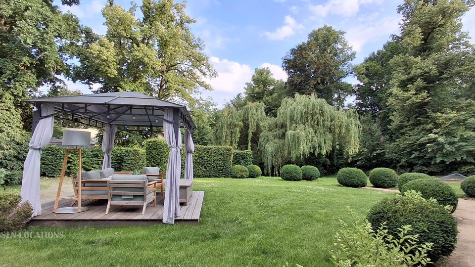 Gepflegter Gartenpark mit großer Rasenfläche, umgeben von hohen Bäumen, Hecken und kugelförmig geschnittenen Sträuchern. Links steht ein offener Pavillon mit Stoffvorhängen und Sitzmöbeln auf einer Holzplattform, rechts verläuft ein sandiger Weg.