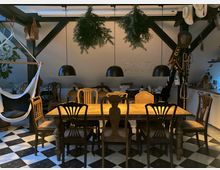 Gemütlicher Essbereich in einem Wohnraum mit klassischem Holztisch, umgeben von unterschiedlichen antiken Stühlen, darunter vier schwarze Hängelampen. Der Raum hat eine Schachbrettfliesen-Bodenoptik, dunkle Dachbalken sowie eine Hängematte und dekorative Gegenstände wie Hüte und Pflanzen.