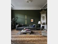 Wohnzimmer mit grüner Wand, klassischer Stuckdecke und Fischgrätenparkett. Zwei graue Sofas, ein heller Teppich, ein niedriger Couchtisch mit Büchern und Kerzen sowie ein dekorativer Kachelofen prägen den Raum.