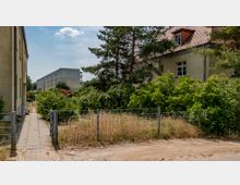 Wohngebiet mit einem schmalen Gehweg, flankiert von einem Metallzaun und dichter Vegetation. Im Hintergrund mehrere Gebäude, darunter ein modernes Wohngebäude und ein Haus mit rotem Ziegeldach.