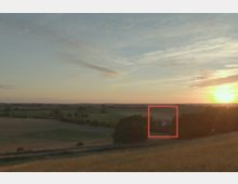 Weite Landschaft mit Feldern und Wiesen, vereinzelt von Bäumen umsäumt. Im Hintergrund ein Sonnenuntergang mit leicht bewölktem Himmel, rechts ein kleines Haus mit rotem Dach inmitten von Bäumen.