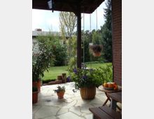 Überdachte Terrasse mit Steinboden, umgeben von Pflanzen in Töpfen, darunter hängende Blumenampeln und ein Holztisch mit Obst. Im Hintergrund ist ein gepflegter Garten mit Rasen, Bäumen und Sträuchern zu sehen.