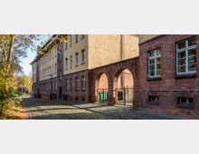 Historisches Gebäude mit roten Ziegelwänden und gelber Putzfassade, verbunden durch eine Arkade aus roten Backsteinbögen, entlang einer mit Kopfsteinpflaster gepflasterten Straße mit Herbstbäumen im Hintergrund.