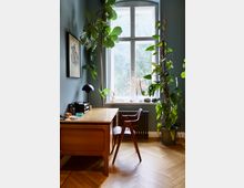 Ein stilvoll eingerichtetes Arbeitszimmer mit einem hölzernen Schreibtisch und Stuhl auf einem Parkettboden. Vor einem großen, weiß gerahmten Fenster stehen mehrere Zimmerpflanzen, die den Raum beleben.