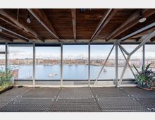 Innenansicht eines modernen Gebäudes mit Metallgitterboden, sichtbaren Holzbalken an der Decke und großen Glasfenstern, die auf einen Fluss und eine Stadtlandschaft mit Wohngebäuden und Booten blicken. Pflanzen in Töpfen stehen links und rechts des Fensters.