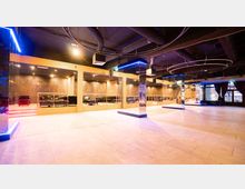 Ein moderner Veranstaltungsraum mit hell beleuchtetem Boden, verspiegelten Säulen und eingelassenen Sitzbereichen mit Sofas, die durch Geländer abgegrenzt sind. Die Decke ist schwarz gestrichen und durch sichtbare Rohre und schlanke Leuchtringe gestaltet, während große Fenster mit Vorhängen seitlich Tageslicht einlassen.