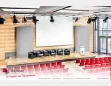Ein moderner Konferenzraum mit einer Bühne, darauf fünf schwarze Stühle, kleine Glastische und ein Rednerpult. Im Hintergrund befinden sich eine große Leinwand und Holzwandpaneele; der Raum ist mit roten Stühlen und großen Fenstern ausgestattet, die Tageslicht hereinlassen.