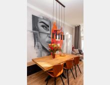 Moderner Essbereich mit großem Holztisch und braunen Lederstühlen unter einer Hängelampe mit mehreren schwarzen Pendelleuchten. An der Wand befindet sich ein großflächiges Graffiti-Porträt, daneben ein Fenster mit Vorhängen sowie ein Heizkörper und ein Sofa in der Ecke.