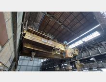 Innenansicht einer großen Industriehalle mit einem massiven gelben Brückenkran. Der Kran verfügt über eine Kabine, Geländer und mehrere Flaschenzüge, die von der Decke hängen; die Halle hat ein hohes Dach mit sichtbarer Metallkonstruktion und Fensteröffnungen, die teilweise Tageslicht hereinlassen.