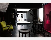 Ein professionelles Fotostudio mit dunklem Hintergrund und heller Beleuchtung. Im Vordergrund sind ein Hocker, ein grüner Stuhl und Beleuchtungs- sowie Kameraausrüstung sichtbar, im Hintergrund steht eine erhöhte weiße Plattform mit Softboxen und Reflektoren.